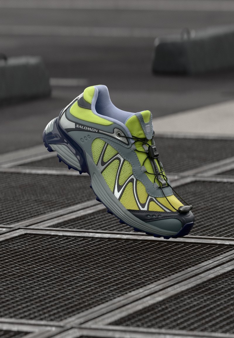 Sapatilha de corrida Salomon com parte superior em malha cinza e amarelo neon, design aerodinâmico, detalhes em preto e um sistema de amarração rápida, apresentando uma sola acolchoada.