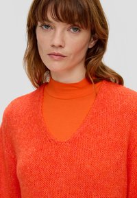 Gebreide oranje trui met een V-hals ontwerp, gelaagd over een aansluitende oranje coltrui. De textuur is zacht met een losse, open weving.