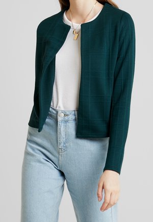 Kobieta ubrana w ciemnozieloną teksturowaną kurtkę z otwartym przodem na białą bluzkę, jasnoniebieskie jeansy z wysokim stanem i warstwowe złote naszyjniki.