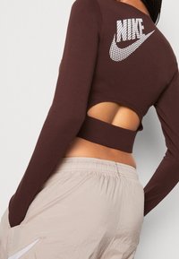 Brun långärmad crop top med utskuret bakstycke, som har en vit genombruten Nike-logga. Matchas med ljusbeige shorts.