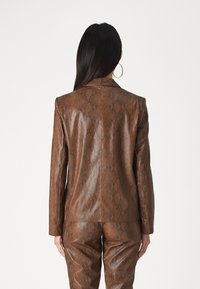 Hosszú sötét hajú nő barna kígyóbőr mintás zakóban és hozzá illő nadrágban áll, háttal nézve, a hátulról látható.