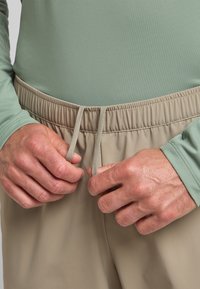 Cintura elastica beige con lacci, abbinata a una camicia a maniche lunghe verde chiaro. Il tessuto appare liscio con una textura sottile.