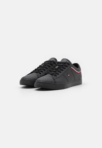 Schwarze Sneaker aus synthetischem Material mit flachen Schnürsenkeln, einer runden Zehenpartie und einem subtilen roten und weißen Akzent entlang der Ferse.