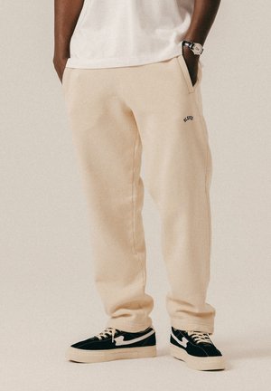 Osoba ubrana w beżowe spodnie dresowe z rękami w kieszeniach, czarno-białe trampki i białą koszulę, stojąca na jednolitym tle.