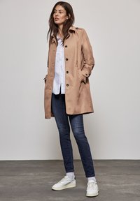 Beige trenchcoat med knäppning, sidofickor och krage, bärs över en vit skjorta och mörkblå jeans, tillsammans med vita sneakers.