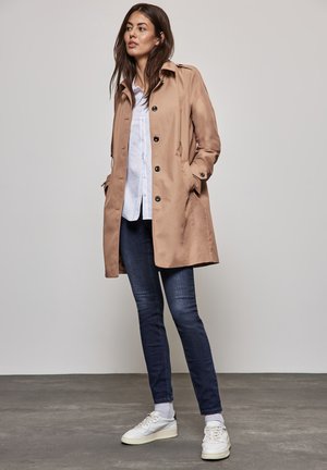 Manteau trench beige avec une fermeture à boutons, des poches latérales et un col, porté sur une chemise blanche et un jean bleu foncé, assorti avec des baskets blanches.