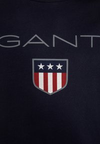 Námořnické tričko s nápisem "GANT" v světle šedé barvě a znakem se třemi hvězdami a červenými a bílými pruhy na spodní části. Materiál: bavlna.