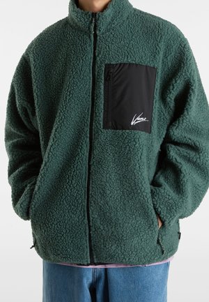 Mann trägt grüne Sherpa-Fleecejacke mit schwarzer Reißverschlusstasche an der Brust und Händen in den Seitentaschen, kombiniert mit blauer Jeans.