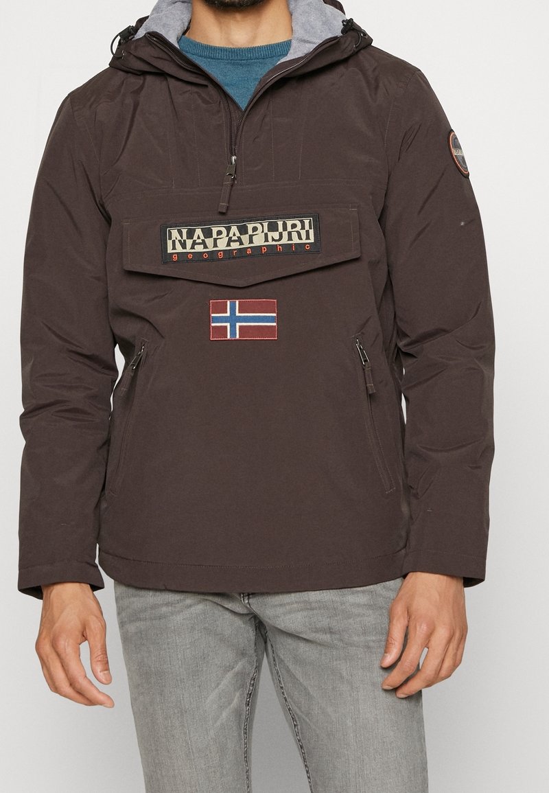 Homme portant une veste Napapijri marron avec une poche à l'avant, un écusson du drapeau norvégien, et un jean gris clair sur un fond uni.
