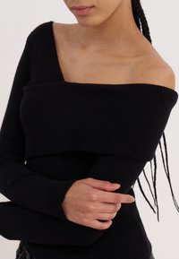 Femme aux cheveux tressés portant un pull noir côtelé à épaules dénudées, les bras croisés sur le torse, sur un fond uni.