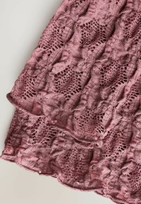 Kanten stof in een gedempte roze tint, met een intricaat bladdesign en schulpachtige randen, die een zachte textuur en delicate ambachtelijkheid demonstreert.