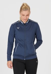Havblå zip-up atletisk jakke lavet af glat stof, med hvide accenter og høj krave. Inkluderer sidelommer og et subtilt logo.