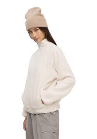 Giacca in fleece beige chiaro con colletto alto, zip frontale e tasche laterali. Texture morbida, vestibilità ampia, abbinata a un berretto lavorato a maglia beige chiaro.
