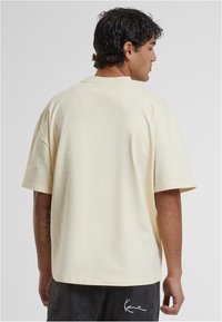 Kreminės spalvos per didelis marškinėlių su trumpomis rankovėmis, lygus paviršius, apvalus iškirpimas ir mažas logotipo detalė ant nugaros krašto.