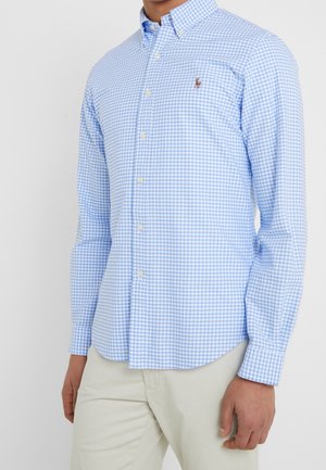 Mężczyzna ubrany w jasnoniebiesko-białą koszulę w kratę z długim rękawem i guzikami z wyszywanym logo, w parze z beżowymi spodniami.