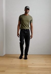 Dark jeans with a tapered fit, olive green t-shirt, and a gray cap. The setting features wooden flooring and a neutral wall.