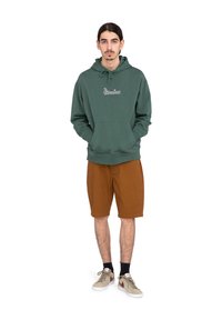 Zelená bavlnená mikina s kapucňou, šnúrkou, prednou kapsou a logom. Hnedé šortky a béžové tenisky s červenými akcentmi. Zobrazený ležérny outfit.