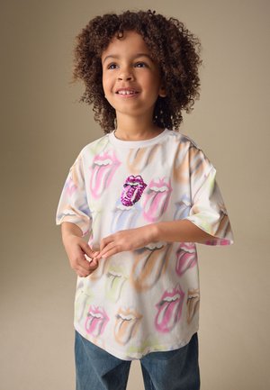 Enfant aux cheveux bouclés souriant, portant un t-shirt blanc oversize avec un motif coloré de lèvres et de langue, et un jean bleu sur un fond uni.
