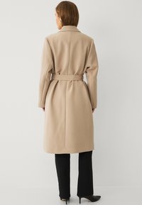 Beige trenchcoat met een riem, voorzien van een kraag en lange mouwen, gemaakt van gladde stof, van achteren weergegeven met een getailleerd ontwerp.