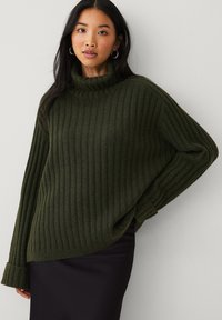 Groen geribbeld sweater met een hoge hals, oversized pasvorm en lange mouwen, gecombineerd met een zwarte rok. Zachte textuur en een ontspannen ontwerp.
