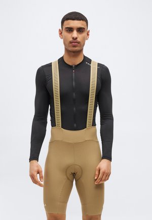 Hombre atlético con una camiseta negra de manga larga con cremallera y pantalones cortos de ciclismo beige con tirantes, de pie contra un fondo liso.