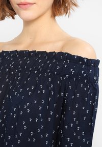 Haut bleu marine à épaules dénudées avec un col froncé, orné de motifs d'ancre blancs. Tissu doux avec une finition texturée.