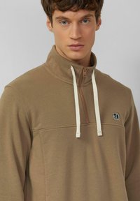 Bruine sweatvest met rits en crèmekleurige trekkoorden, voorzien van een halve ritssluiting bij de kraag en een klein logo op de borst. Zachte stof met een gestructureerde afwerking.