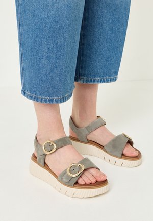Fødder med grå ruskindssandaler med store gyldne spænder og hvide platformsåler, kombineret med korte blå cowboybukser.