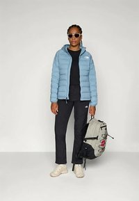 Sieviete, kas valkā gaiši zilu pūkainu jaku, melnas bikses, bēšus sporta apavus, saulesbrilles, tur pelēku un melnu mugursomu uz vienkārša gaiša fona.