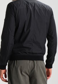 Svart bomberjacka i ett smidigt, lätt material som har ribbade mudd och nederkant samt en enkel, strömlinjeformad design.