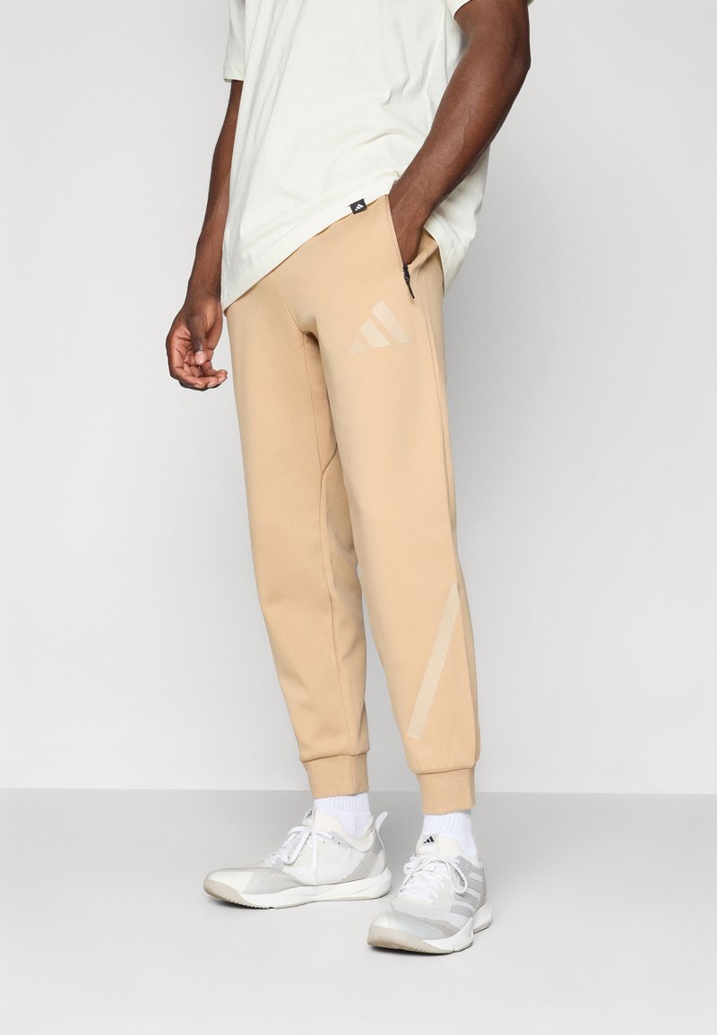 Calças jogger beje feitas de tecido macio, com bolsos laterais, um design afunilado e três riscas diagonais na mesma cor na perna.