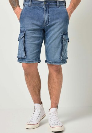 Mann trägt blaue Denim-Cargoshorts mit Klappentaschen und weißen High-Top-Sneakern, steht mit den Händen in den Taschen vor einem einfarbigen Hintergrund.