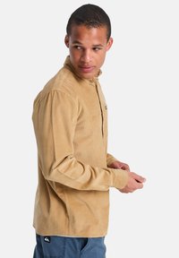 Μπεζ πουκάμισο από κορδόνι με κουμπιά, γιακά τύπου spread, πλαϊνές σκισμές και λογότυπο σε μπαλώματα στην τσέπη, συνδυασμένο με μπλε παντελόνι.