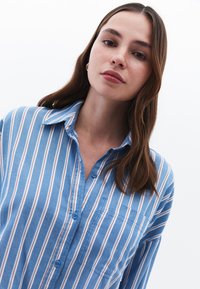 Camicia di cotone azzurro chiaro a righe con strisce rosse e bianche verticali, colletto, frontale con bottoni e una tasca sul petto. Tessuto morbido.