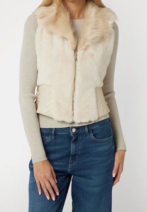 Frau trägt beige Kunstpelz-Weste mit gesteppter Seitenpartie über beigem, geripptem Langarmshirt und blauer Jeans, Hand ruht auf dem Oberschenkel.