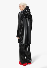 Abrigo negro, brillante y a prueba de agua con capucha, mangas largas y un ajuste holgado, combinado con pantalones negros a juego y calzado rojo brillante.