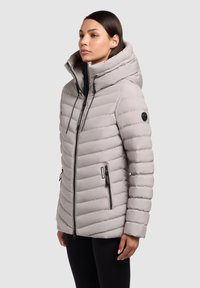 Lys grå pufferjakke med høj krave, lynlås foran og to sidelommer. Har vandret quiltning og et logo-patch på ærmet.