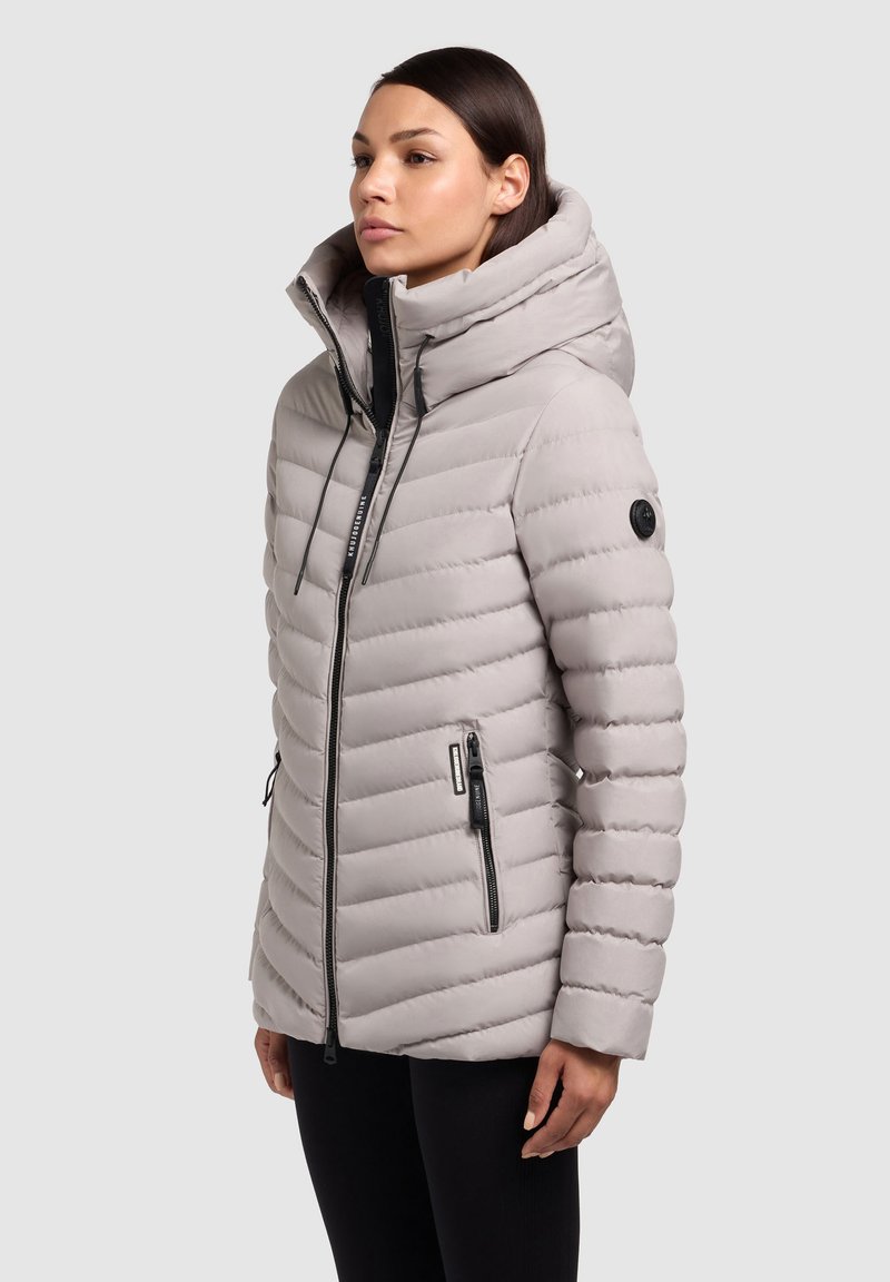 Lys grå pufferjakke med høj krave, lynlås foran og to sidelommer. Har vandret quiltning og et logo-patch på ærmet.