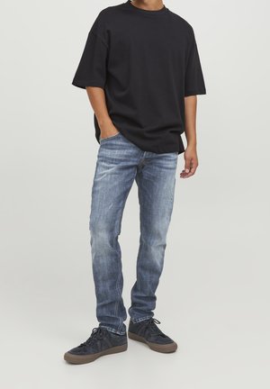 Person, die ein lockeres schwarzes T-Shirt, ausgewaschene blaue Jeans und schwarze Sneakers trägt, steht mit einer Hand in der Tasche vor einem schlichten Hintergrund.