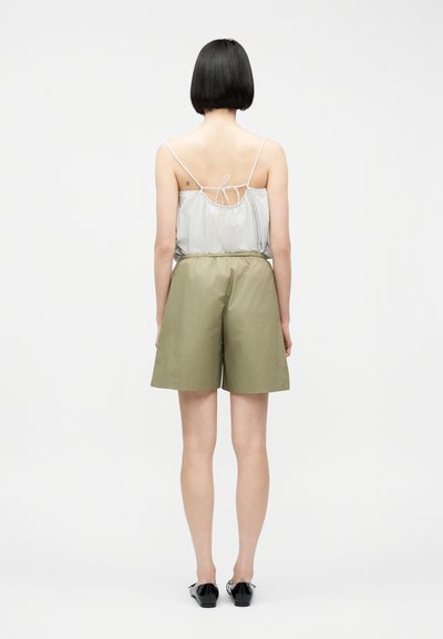 Γυναίκα με κοντά μαύρα μαλλιά, ντυμένη με ριγέ αμάνικη μπλούζα με λεπτές τιράντες και φαρδιά χακί βερμούδα, στέκεται γυρισμένη από την πλάτη, σε λευκό φόντο.