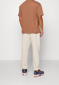Mann, der mit dem Rücken steht, trägt ein braunes kurzärmliges Shirt, cremefarbene Jogginghosen und marineblaue Sneakers mit orangefarbenen Akzenten.