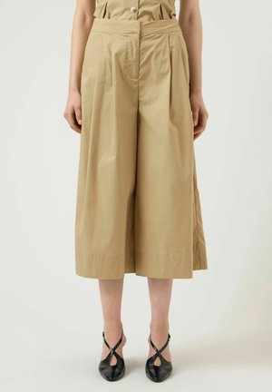 Donna che indossa pantaloni beige larghi e corti e tacchi neri con punta appuntita e cinghie incrociate, in piedi davanti a uno sfondo uniforme.
