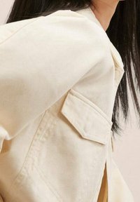 Beżowa bawełniana kurtka z zaostrzoną kieszenią, teksturowaną powierzchnią i luźnym krojem, z subtelnym połyskiem i widocznymi szczegółami szwów.