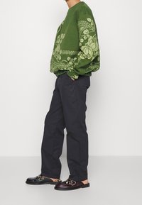 Personne portant un pull vert à motif floral, un pantalon noir droit et des mocassins en cuir dépareillés avec des détails métalliques à boucle.
