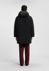 Giacca a vento nera con un grande cappuccio, caratterizzata da una texture liscia e una vestibilità comoda, indossata sopra pantaloni bordeaux e scarpe nere.