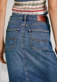 Jeansskørt med lige snit, mørkeblå farve, synlige sting, to baglommer og et brunt lædermærke i taljen.
