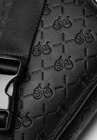 Sac en cuir noir texturé avec des motifs de bicyclettes embossés, un motif en croix et une sangle robuste avec un accent en boucle métallique.