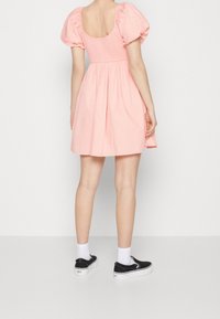 Vestido corto rosa claro con mangas abullonadas, cuerpo fruncido y falda acampanada. Llevado con zapatos negros de tipo slip-on y calcetines blancos por el tobillo.