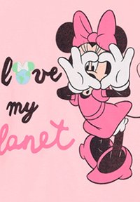 Camiseta rosa con un personaje de dibujo animado formando un corazón con las manos. El texto dice "Amo mi planeta". Se aprecian detalles en negro y verde.