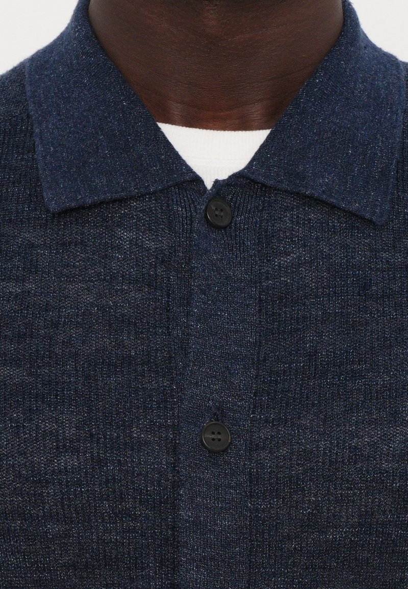 Dark blue knit shirt with a buttoned front, featuring a short collar and a textured pattern. Two black buttons visible near the neck.
